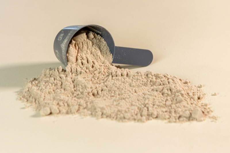 Tipped scoop spilling beige protein powder on a cream surface
